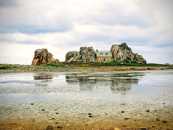 Un grand bâtiment isolé, adossé à un relief rocheux, situé sur une plage de sable avec une étendue d’eau calme en premier plan.