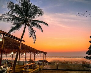 A beachside restaurant with outdoor seating, a palm tree, and a view of the ocean at sunset. - Nalanda Retreat Goa (Mandrem)