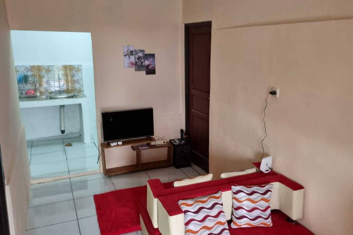 A living room with a TV, a wooden TV stand, a red sofa, and a red rug. There is a door leading to another room with a tiled floor and a window.