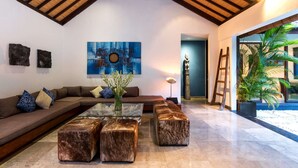 A modern living room with a high ceiling, a large abstract painting, a sofa with patterned cushions, a glass coffee table, and a view of a tropical garden through a glass door. - Rent a Luxury Villa in Bali Close to the Beach, Bali Villa 2016 (Seminyak)