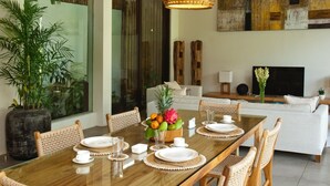 Un coin repas avec une table en bois dressée pour quatre personnes, entourée de chaises en osier. Un panier de fruits contenant différentes sortes de fruits est posé sur la table.