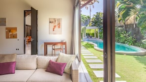 Sala de estar com sofá e mesa, um quadro na parede e vista para uma piscina e jardim.