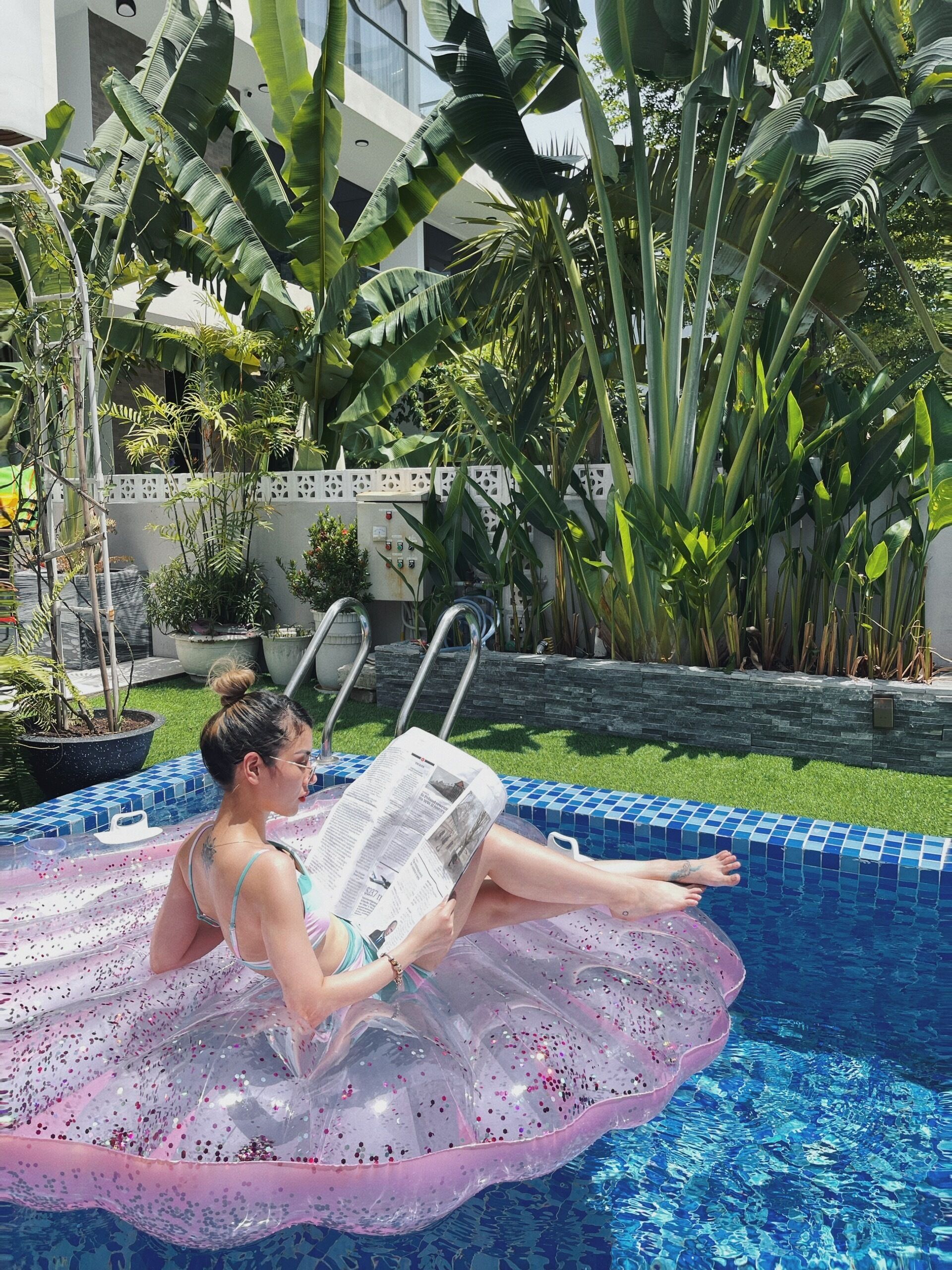 A person relaxing in a pool with a transparent inflatable ring, surrounded by lush greenery and a modern building in the background.