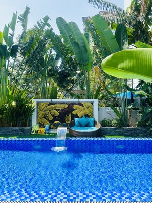 A swimming pool with a blue mosaic border, surrounded by lush tropical plants and a decorative wall with a banana leaf mural. - MAY CHILL HOUSE VILLA (Da Nang)