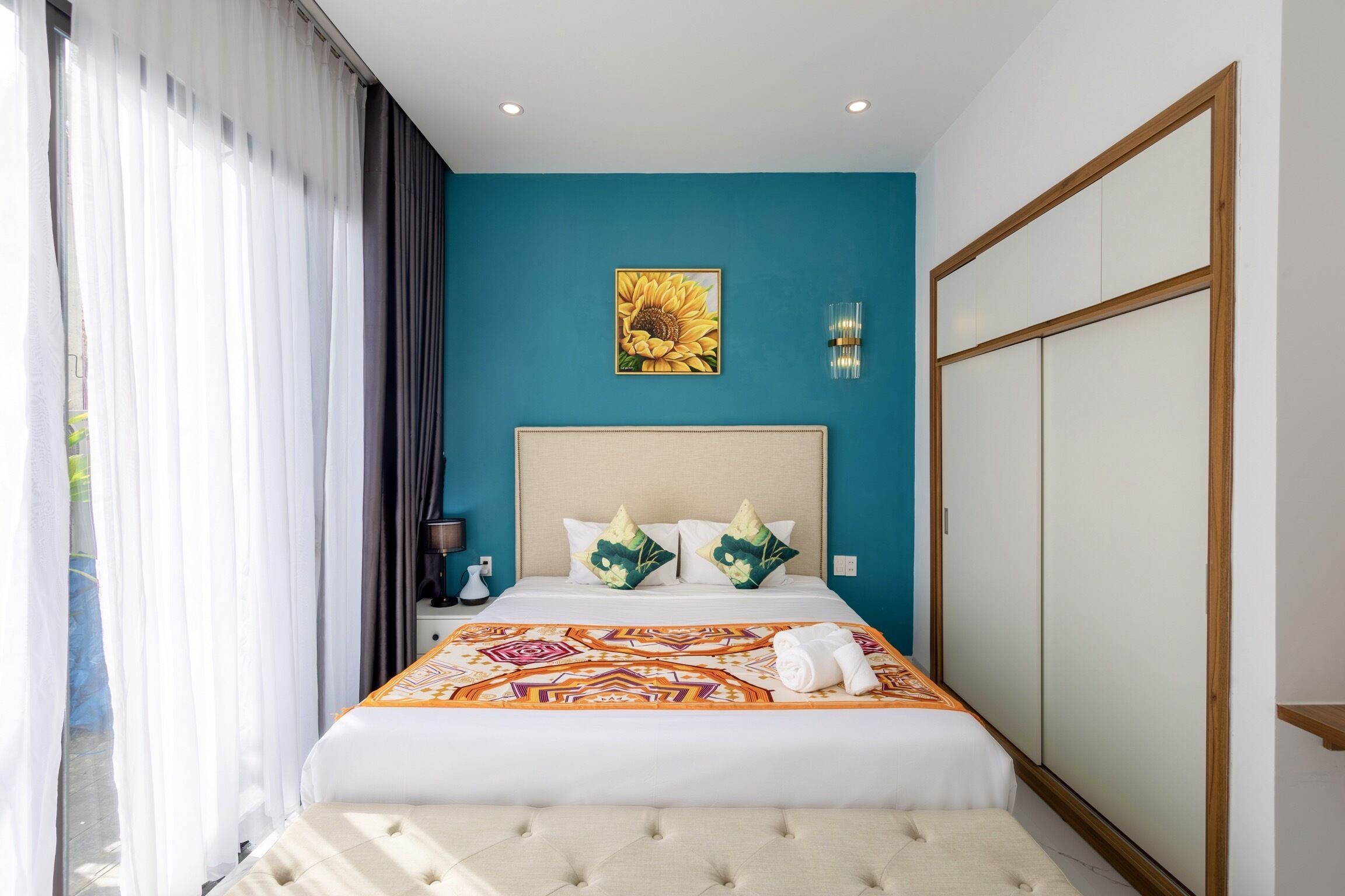 Ein Hotelzimmer mit einem großen Bett, einer blauen Wand als Blickfang, einem Sonnenblumenbild und einem Kleiderschrank.
