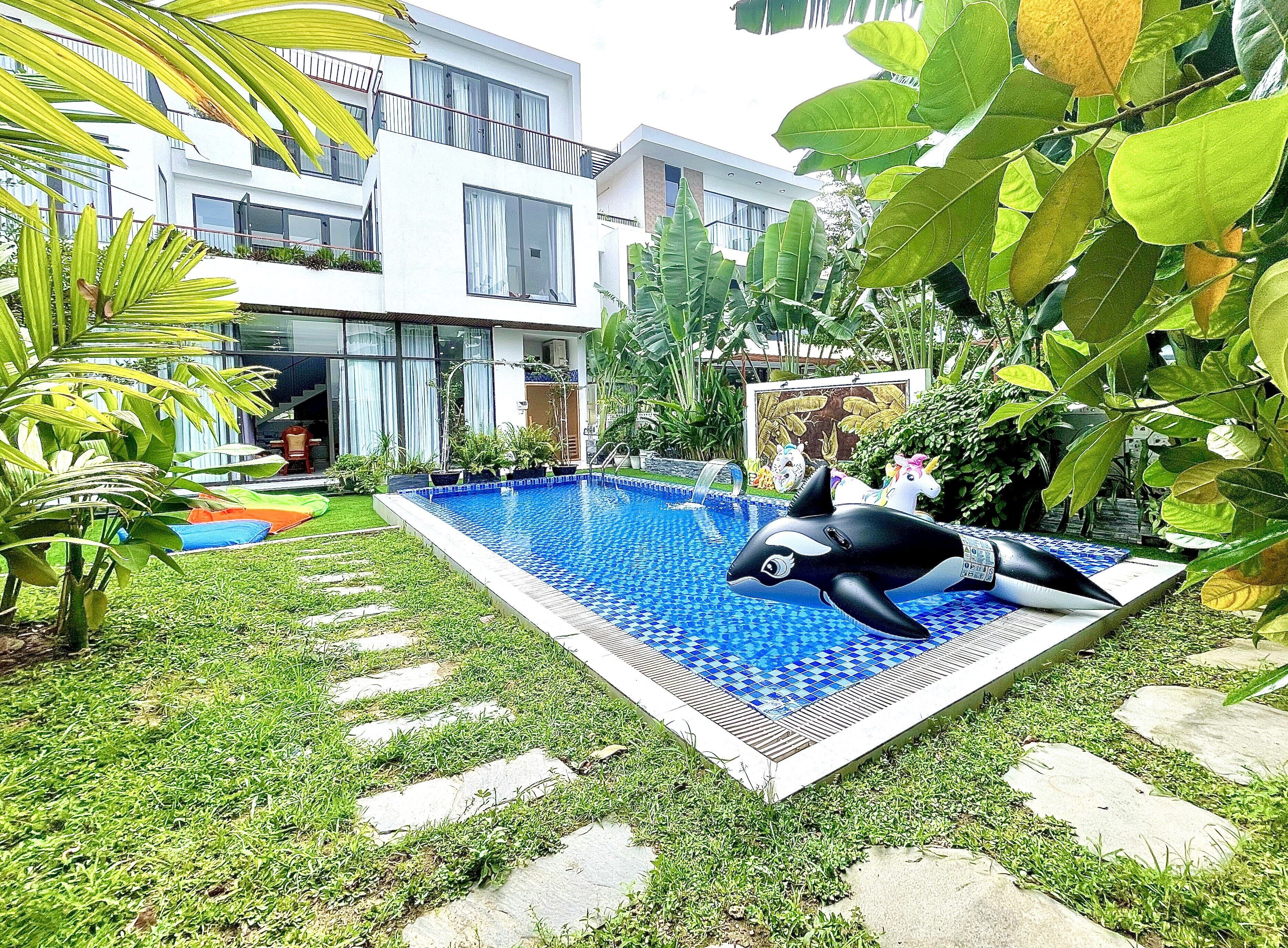 A swimming pool with a dolphin float, surrounded by lush greenery and a modern building in the background.