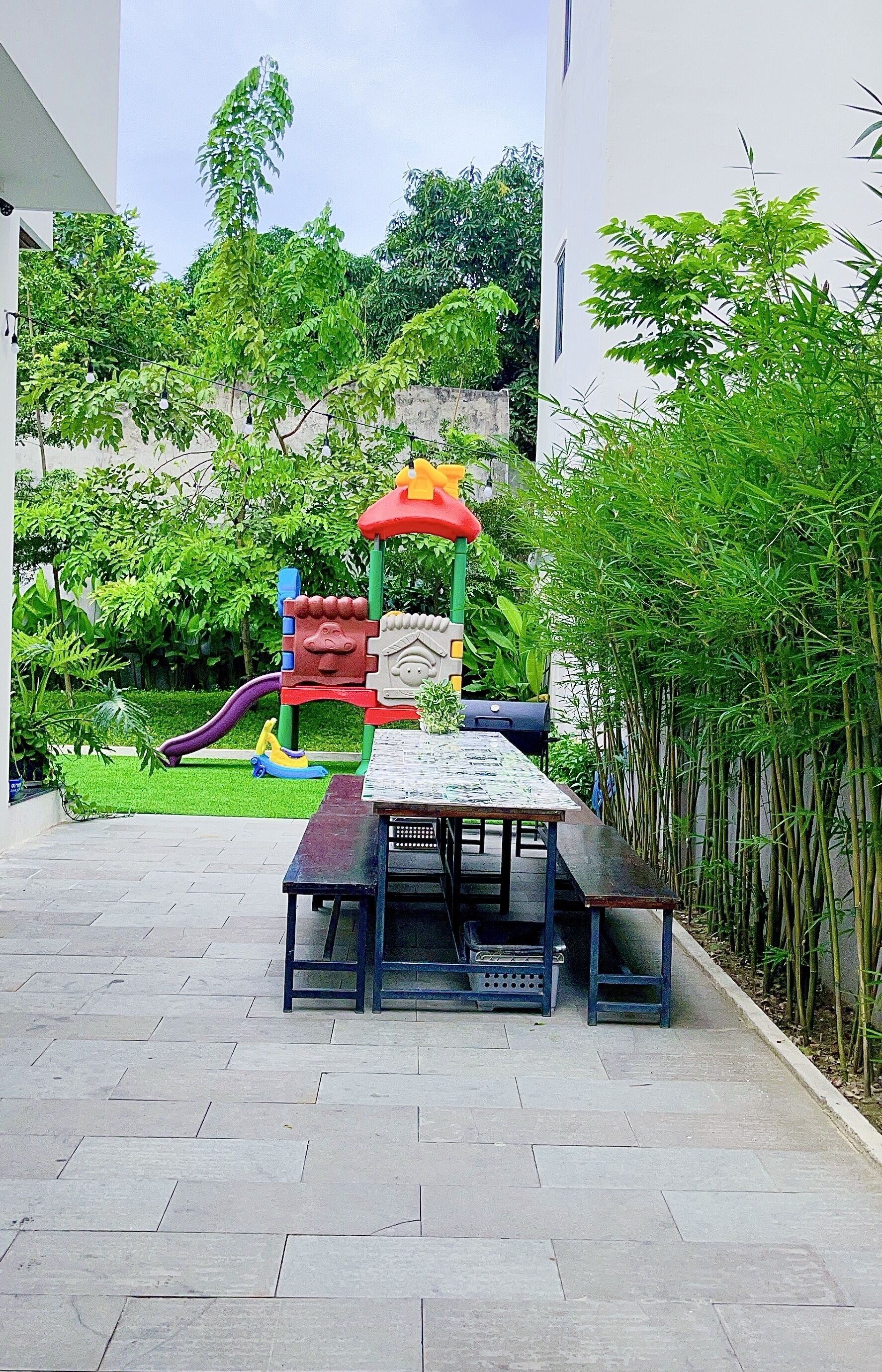 Zona de juegos infantiles con tobogán y estructuras para escalar, rodeada de vegetación y un patio empedrado.