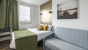 Un dormitorio con una cama, una manta verde, un sofá azul con estampado, una ventana con cortinas y un cuadro colgado en la pared.
