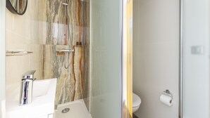 Baño con pared de ducha de mármol, ducha con cerramiento de vidrio, lavamanos blanco con accesorios cromados y un inodoro con dispensador de toallas de papel.