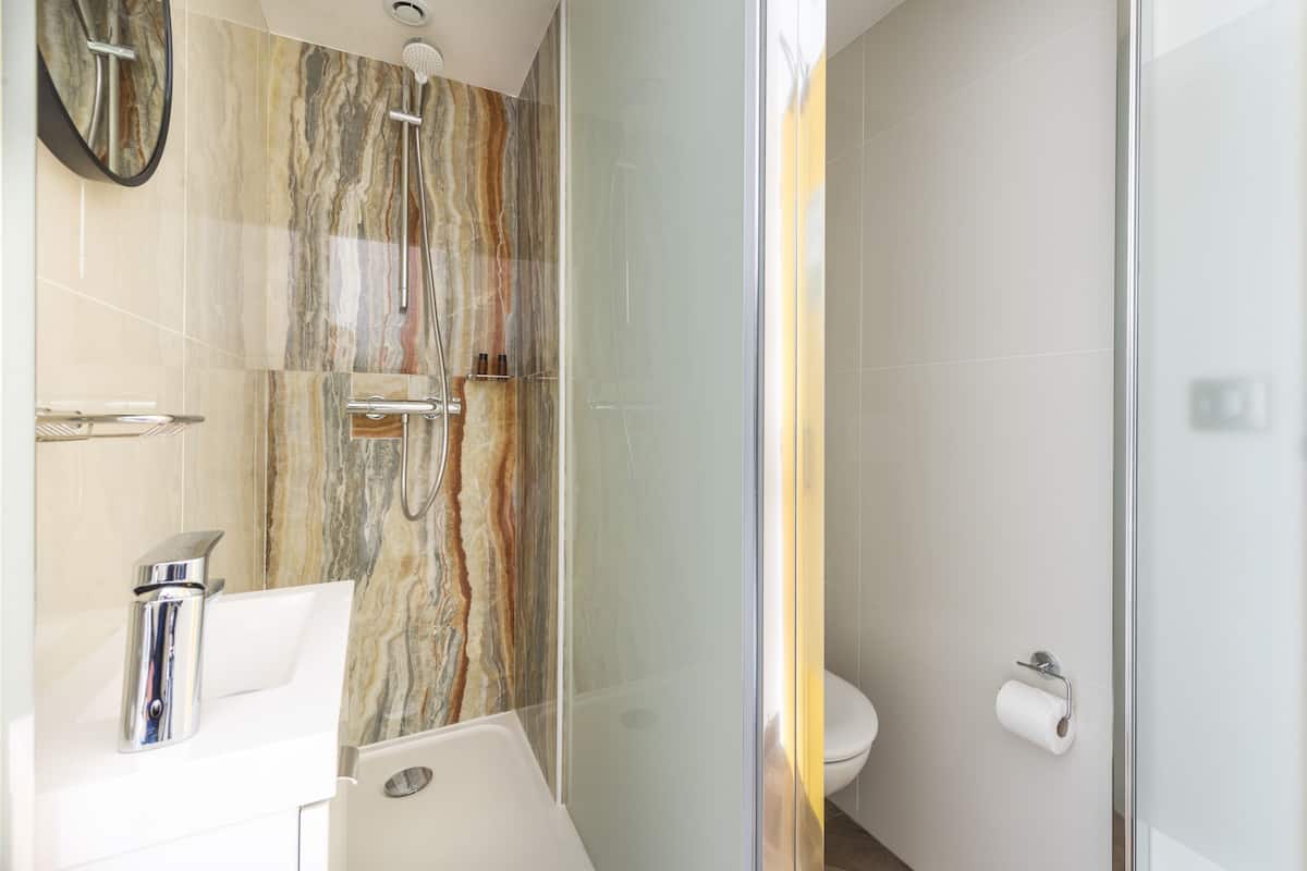 Une salle de bain avec un mur de douche en marbre, une douche fermée par des parois de verre, un lavabo blanc avec des robinets chromés et des toilettes dotées d’un porte-serviettes en papier.