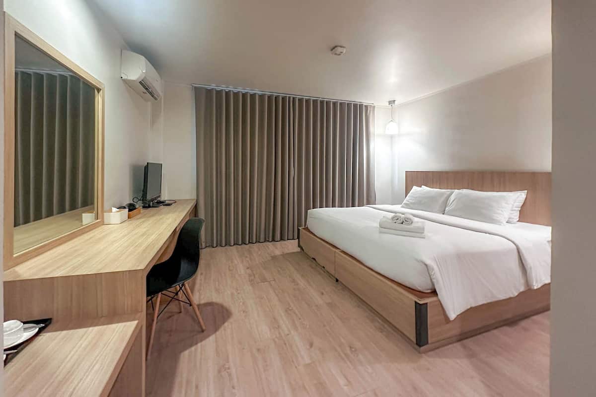 Une chambre d’hôtel avec un bureau en bois, un lit avec une literie blanche, un grand miroir et une fenêtre avec des stores verticaux.
