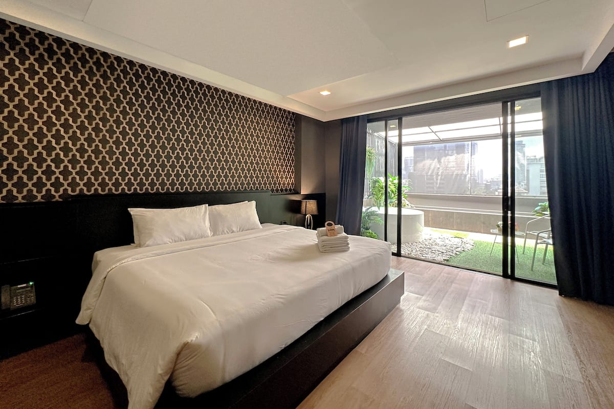 Une chambre d’hôtel moderne avec un grand lit, un mur orné de motifs, une vue à travers une porte coulissante et une petite table avec une lampe.