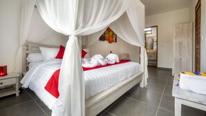 Ein Himmelsbett mit weißer Bettwäsche und roten Akzenten, ein Nachttisch mit roter Lampe und Blick ins Badezimmer durch eine geöffnete Tür.