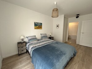 A bedroom with a bed, two bedside tables, a hanging light fixture, a door, and a small painting on the wall.