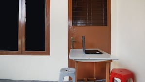 Ein Raum mit Holztür, einem Fenster mit Metallgitterstäben, einem kleinen Tisch mit einem Laptop, einem grauen Hocker und einem roten Hocker.
