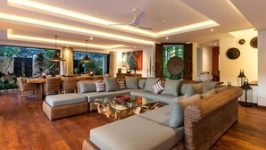 A spacious living area with a large sofa, a glass coffee table, and wicker furniture. There is a dining area with wicker chairs and a table, and a ceiling fan. The room has large windows and a view of greenery outside. - Modern 4BR Villa W/ Pool & Balcony, Bali Villa 2220 (Kerobokan)