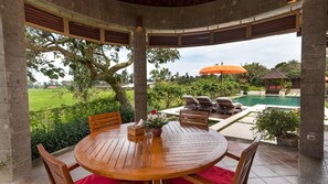 A covered outdoor seating area with a wooden table and chairs, overlooking a pool and lush greenery. - Modern 3BR Villa W/ Pool Area & Garden, Bali Villa 2232 (Sukawati)