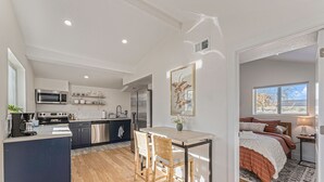 Une cuisine moderne dotée d’appareils électroménagers en acier inoxydable, un coin repas avec un îlot central et des chaises en osier, et une chambre à coucher avec un grand lit et une vue sur le ciel par une fenêtre.