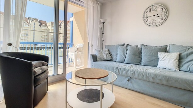 A modern living room with a sofa, a round coffee table, and a large window offering a view of buildings. - Bright Apartment Near Beach with Parking, Balcony & Modern Amenities – Stella (Cucq-Stella Plage)