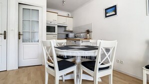 A compact kitchen with white cabinets, a microwave, and a stove. There is a dining table with white chairs and black cushions. - Bright Apartment Near Beach with Parking, Balcony & Modern Amenities – Stella (Cucq-Stella Plage)
