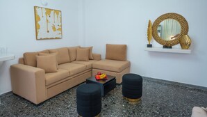 Een woonkamer met een beige bank, zwarte poefjes, een bijzettafeltje met fruit, een spiegel met gouden lijst en een abstract kunstwerk met gouden details.