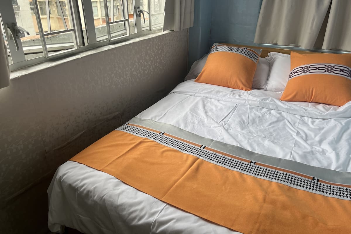 A bed with white and orange bedding, a patterned pillow, and a view of buildings through a window.