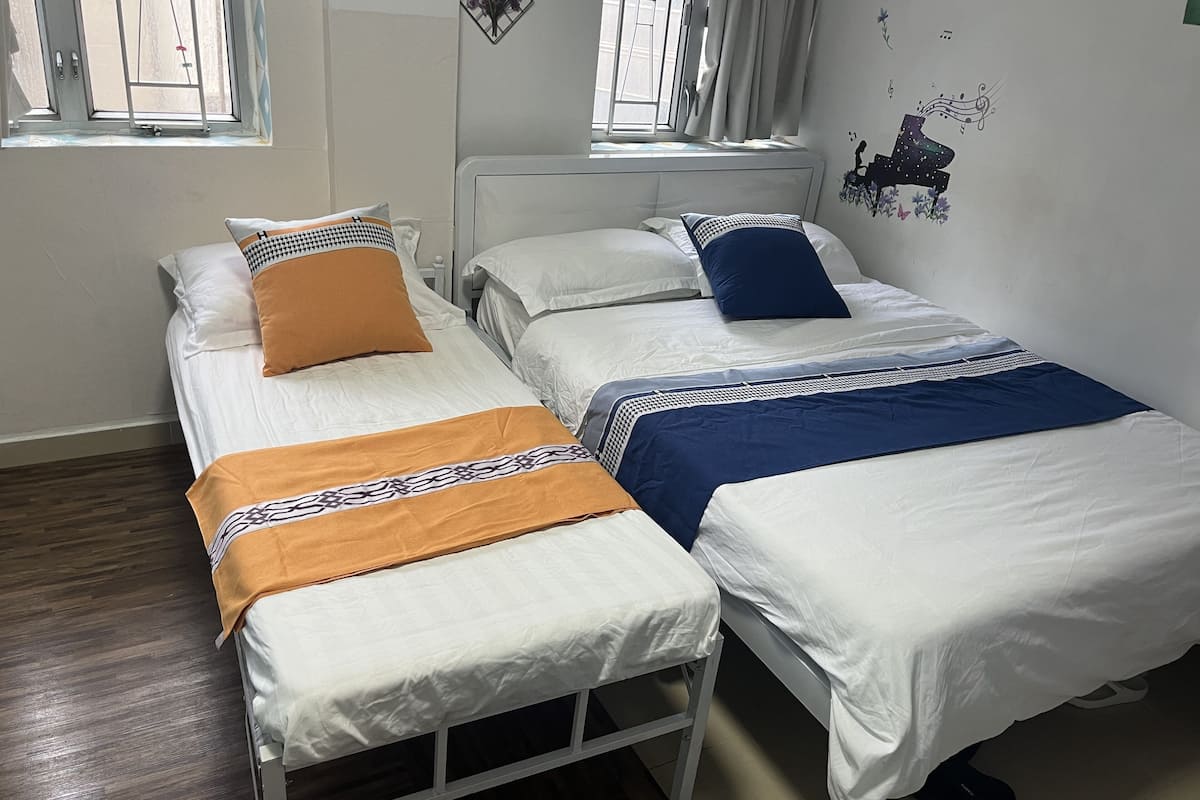A twin room with two beds, each with white bedding and patterned pillows. There are windows with curtains, a radiator, and wall decorations including a musical note and a flower.