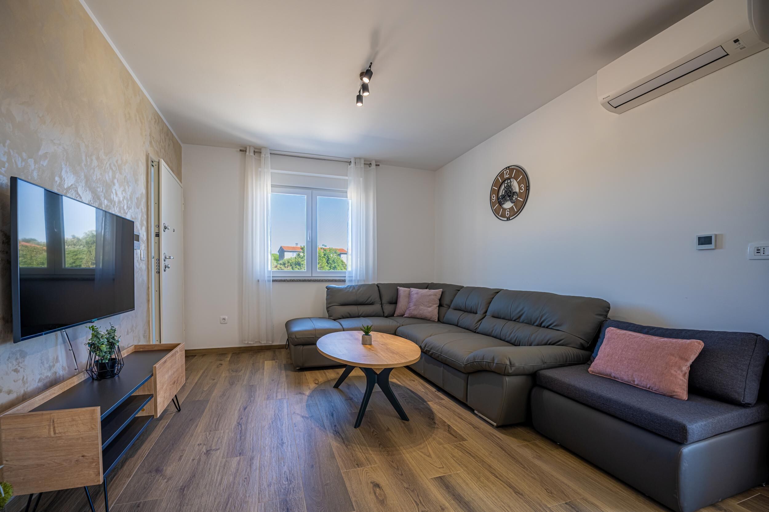 A modern living room with a sectional sofa, a flat-screen TV, a wooden coffee table, and a large window.