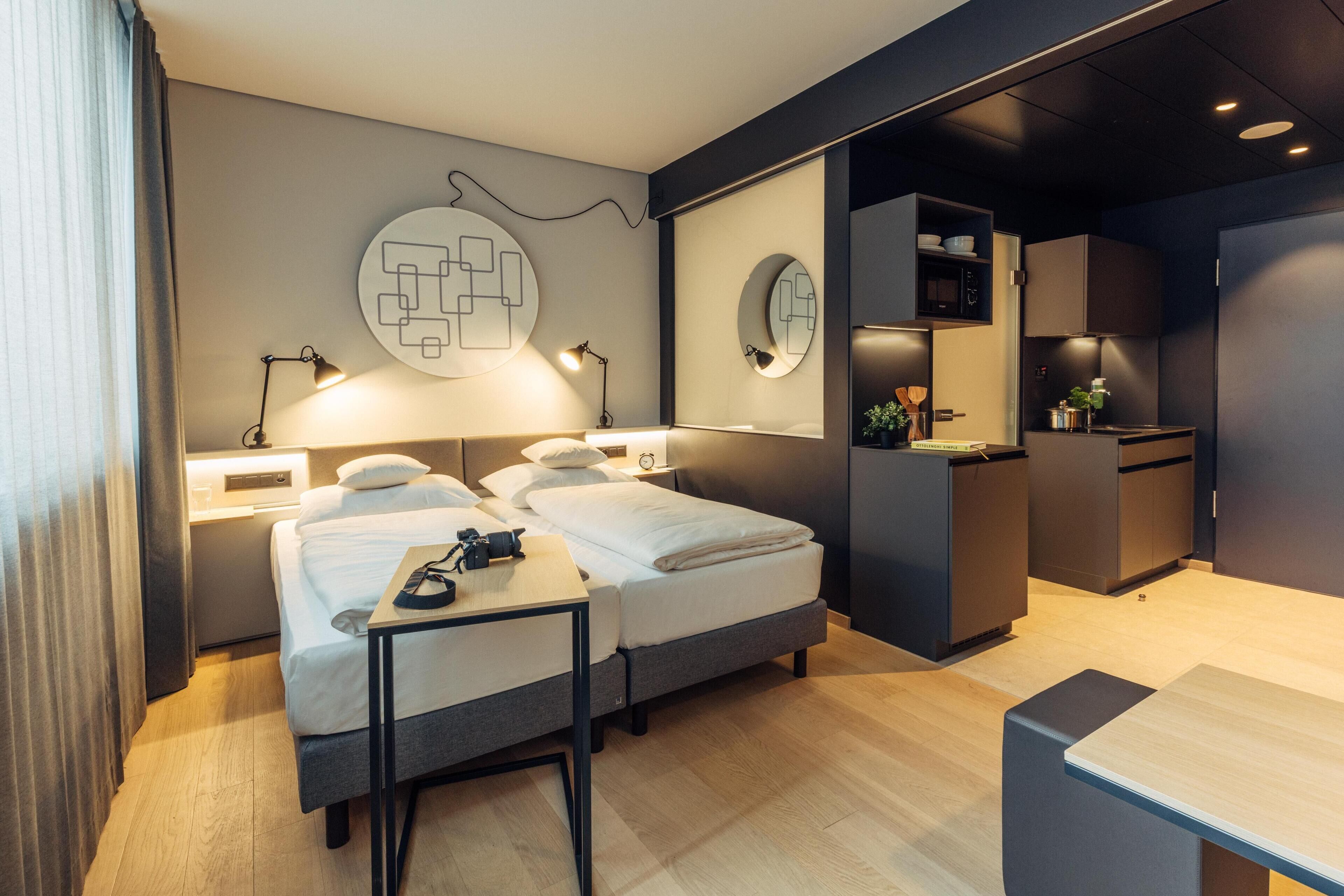 Una habitación de hotel moderna con una cama doble, mesitas de noche, un escritorio, un espejo y una kitchenette.