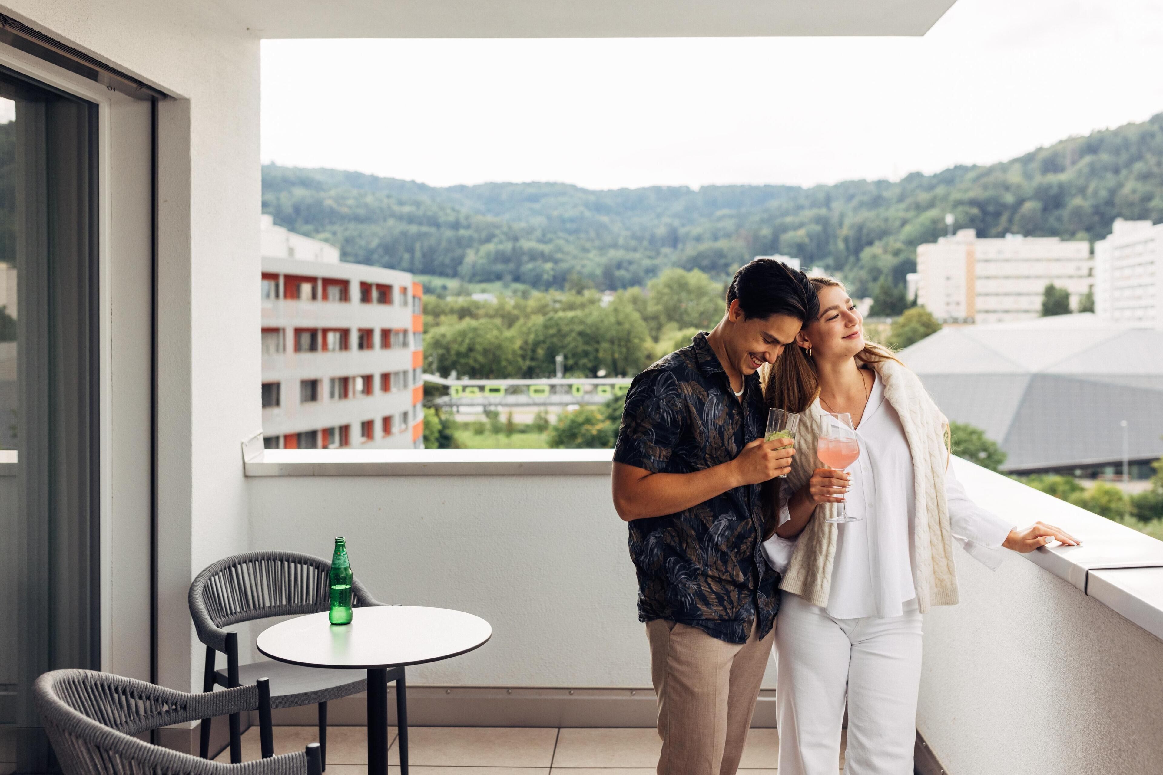 A balcony with a view of buildings and greenery. There is a table with a bottle and two chairs.