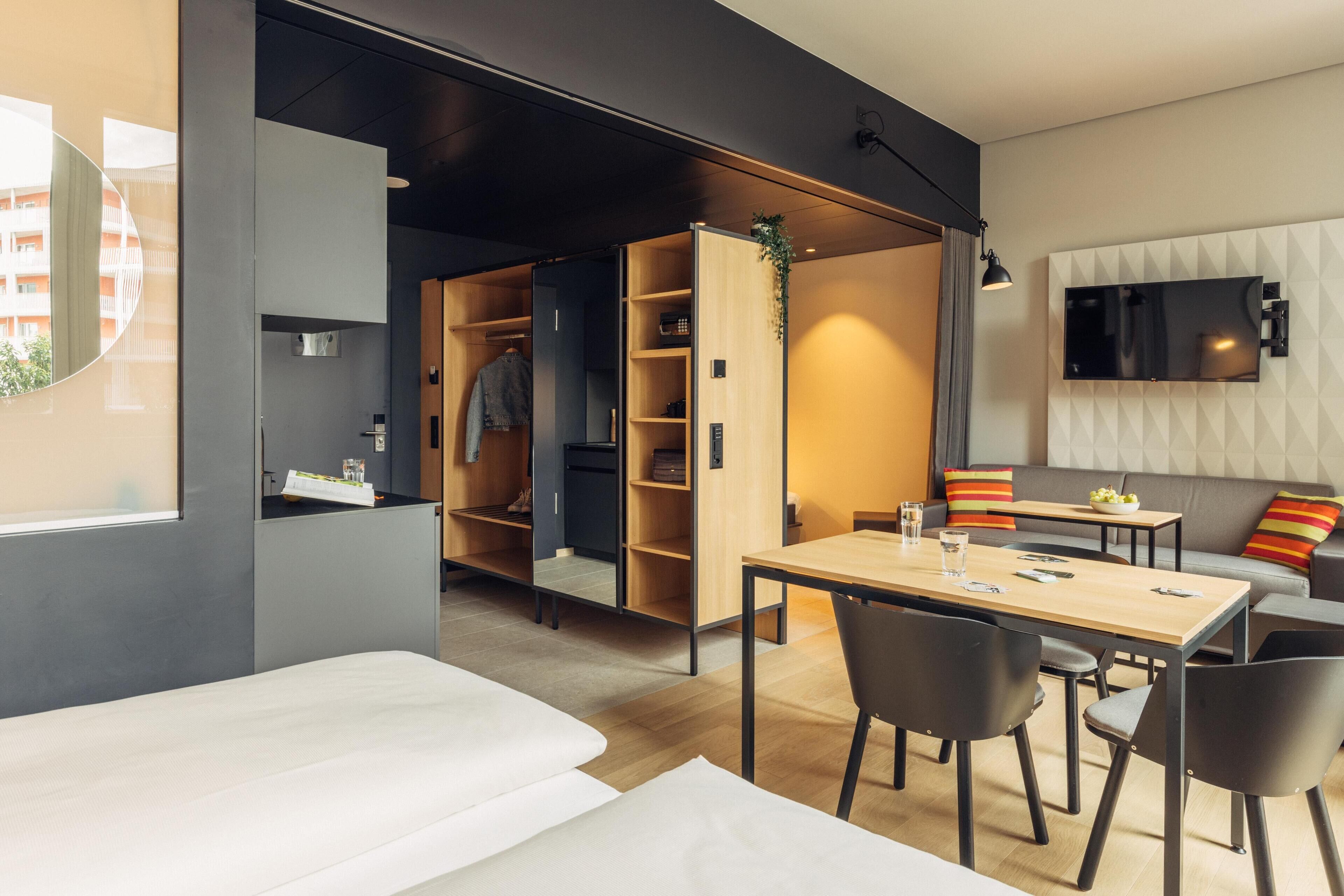 A modern hotel room with a bed, a dining area with a table and chairs, a built-in wardrobe, a TV mounted on the wall, and a window with a view of buildings.