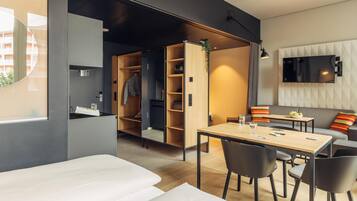 A modern hotel room with a bed, a dining area with a table and chairs, a built-in wardrobe, a TV mounted on the wall, and a window with a view of buildings.