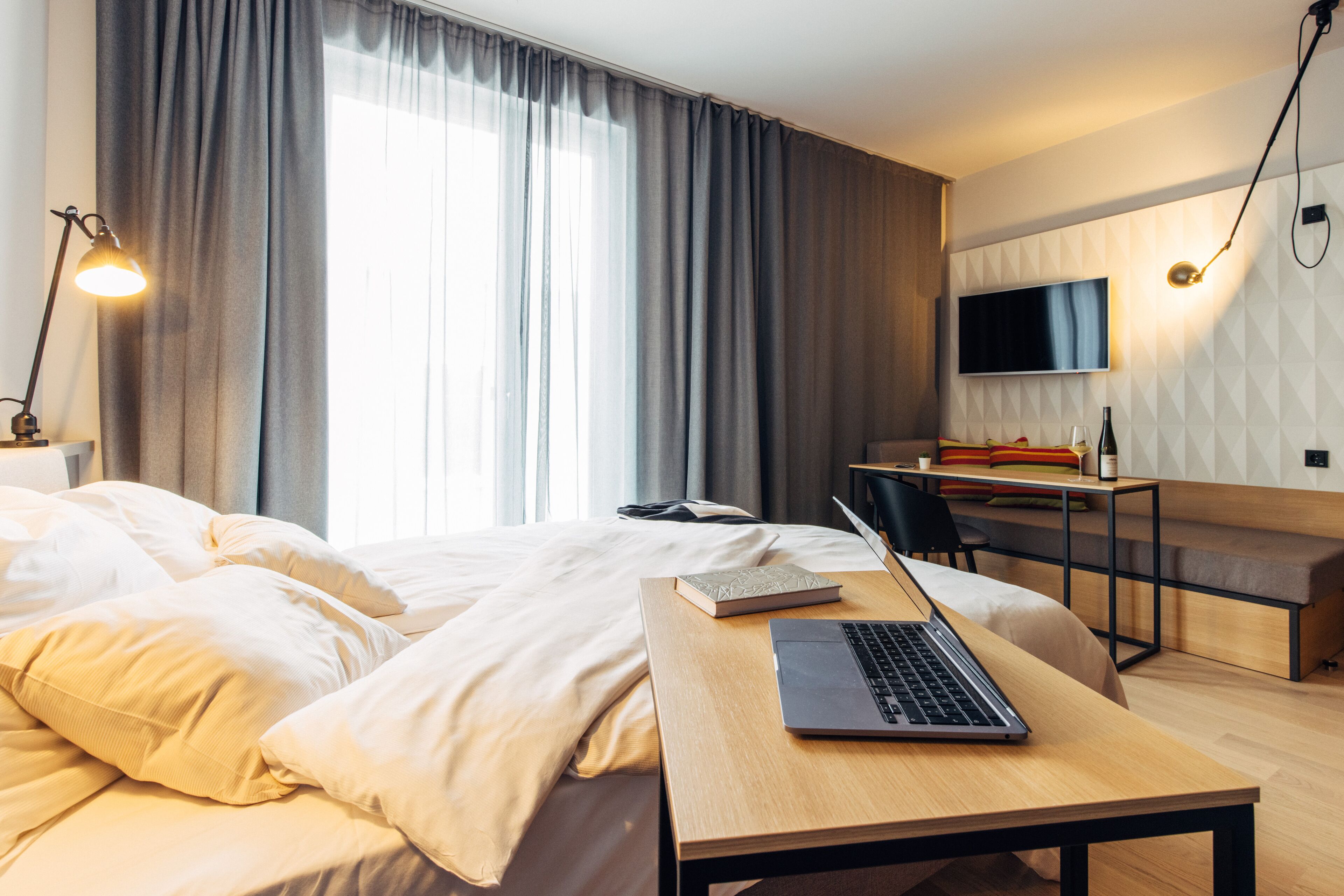 A modern hotel room with a bed, a desk with a laptop, a TV, and a seating area.