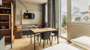 Habitación de hotel moderna con sofá, mesa de comedor y sillas, televisor montado en la pared y un ventanal con vistas a la ciudad.