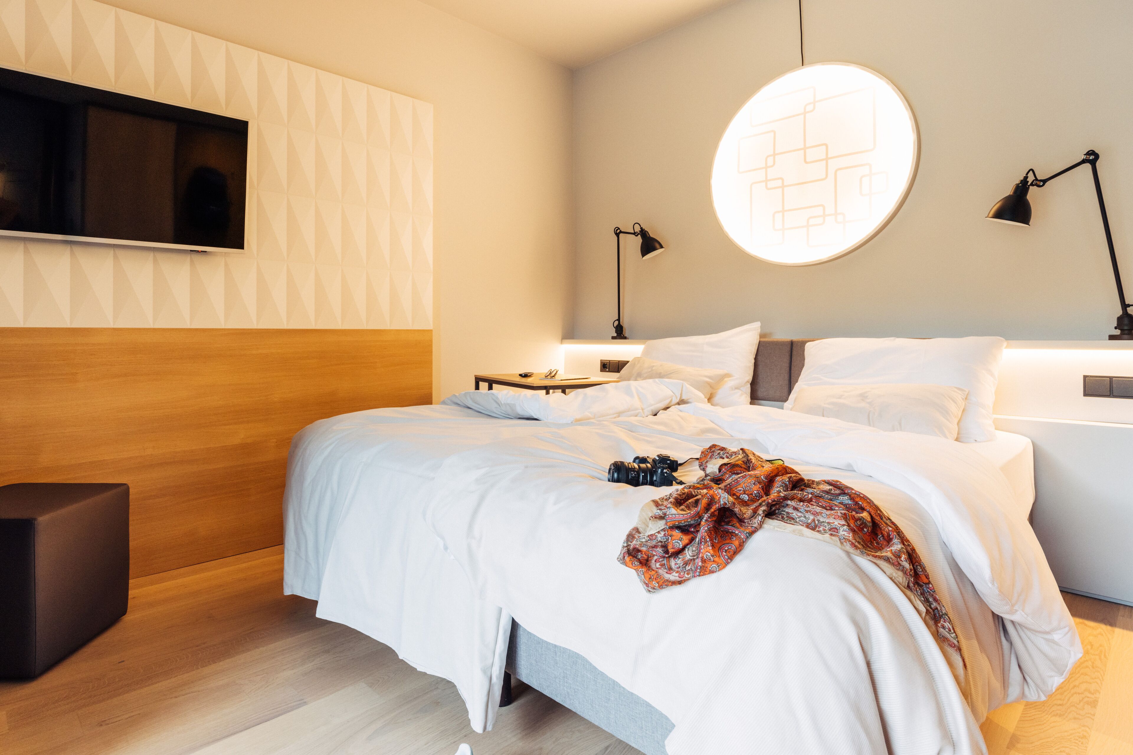 Ein modernes Schlafzimmer mit einem Holz-Kopfteil, einem großen Bett mit weißer Bettwäsche, einer runden Deckenleuchte und einem angebrachten Fernseher.