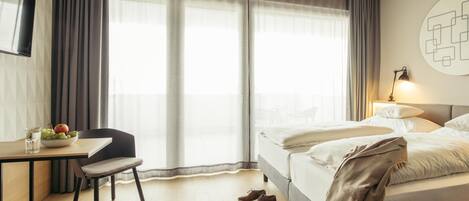 Une chambre d’hôtel comprenant un lit, un bureau avec une chaise, un téléviseur fixé au mur et une grande fenêtre avec des rideaux.