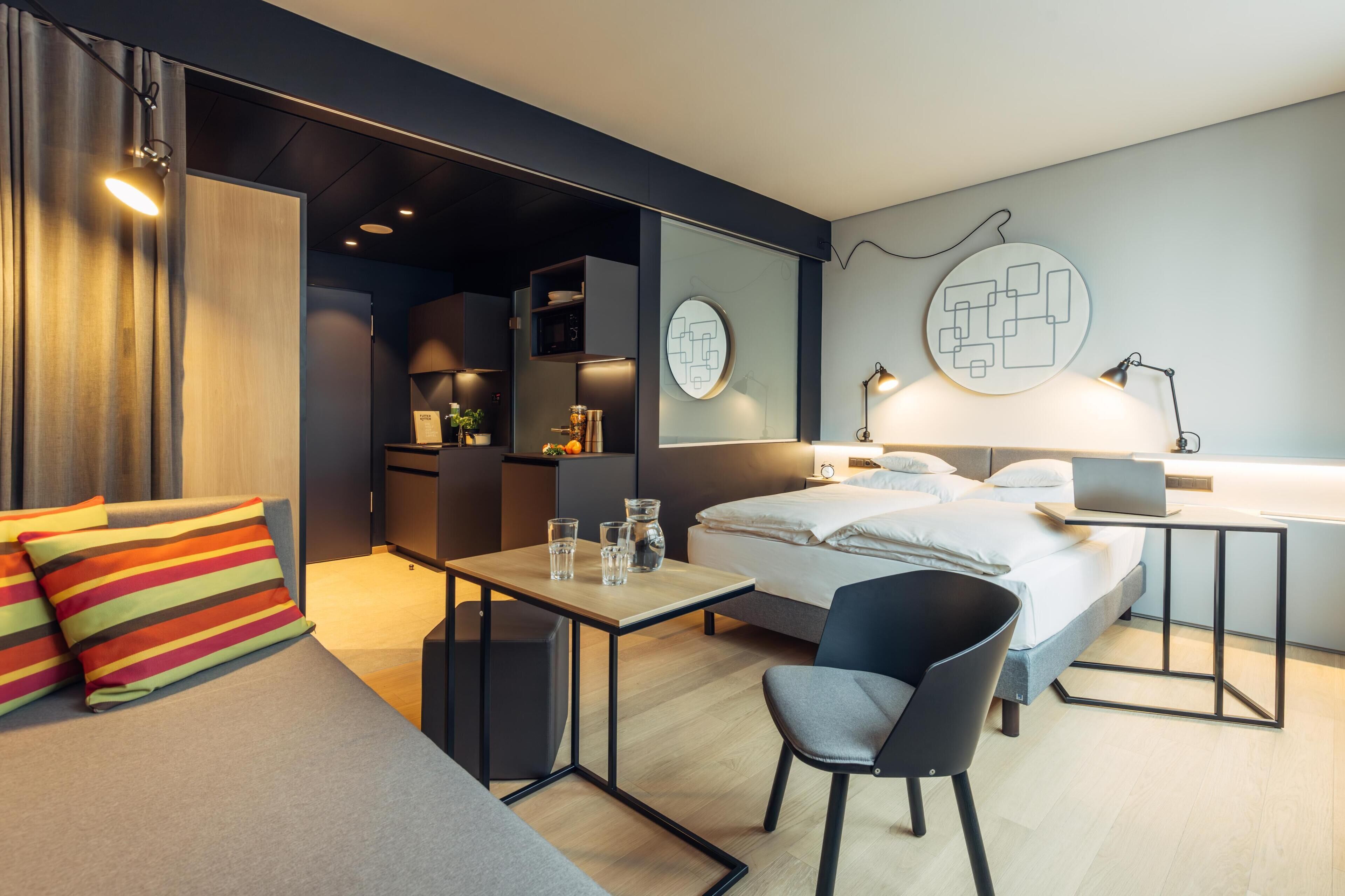 A modern hotel room with a bed, a desk, a chair, a small table, and a sofa.