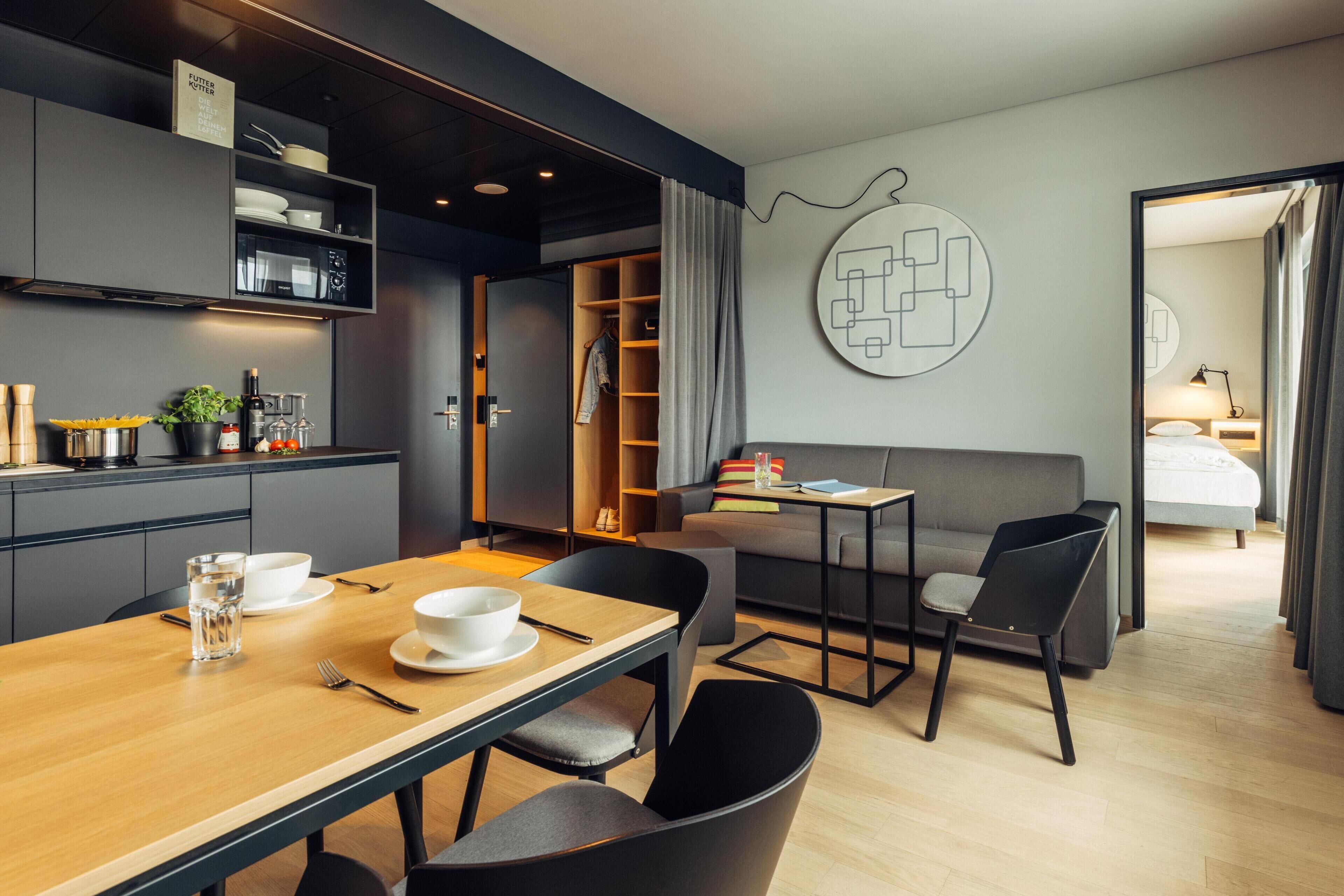 Una habitación de hotel moderna con zona de comedor, sofá, mesita y kitchenette.