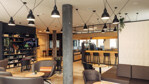 Un espacio lounge moderno con diversas opciones de asiento, incluyendo sofás y sillones, y un pilar central. Hay mesas con revistas y pequeñas plantas.