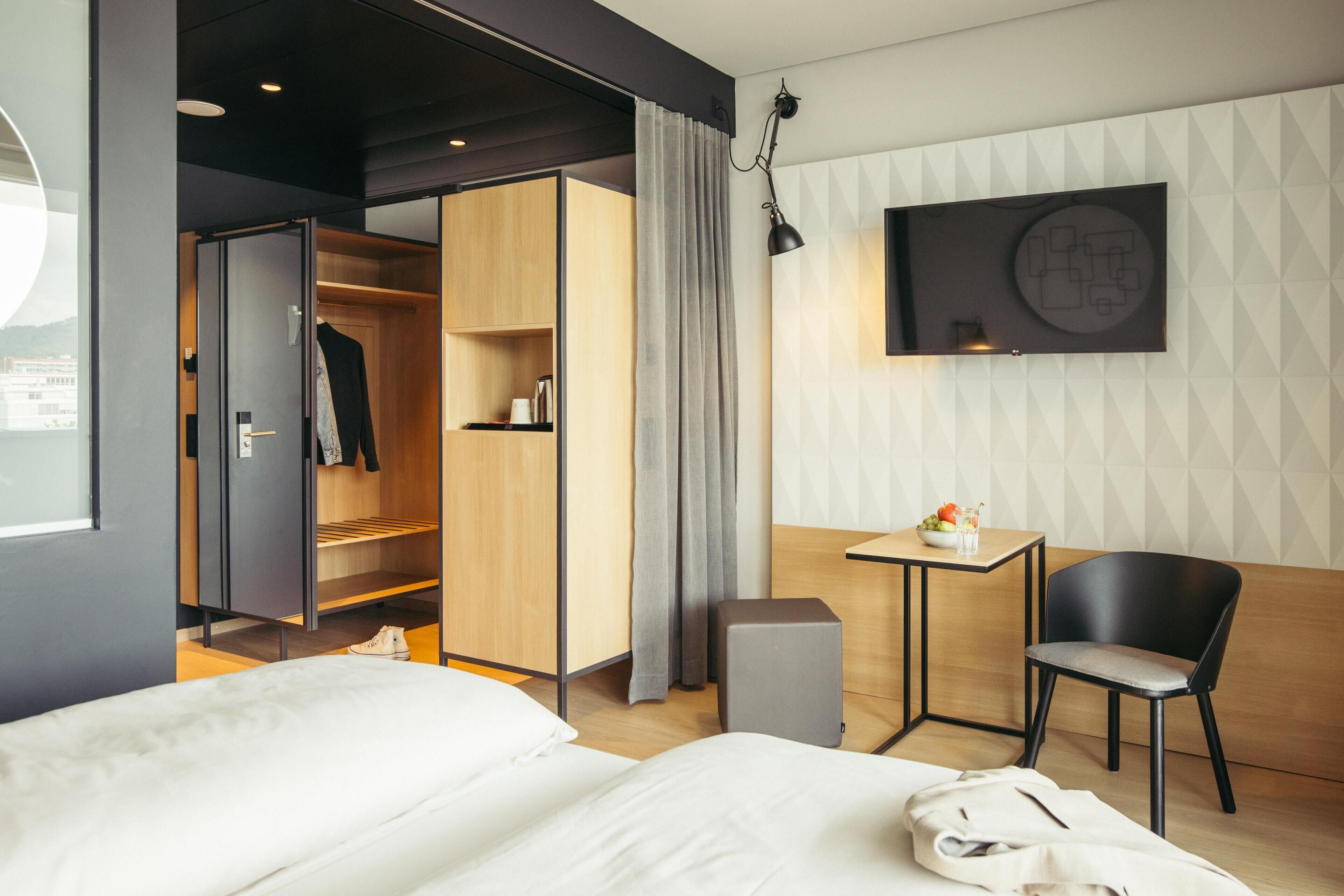 A modern hotel room with a bed, a wardrobe, a desk with a chair, and a TV mounted on the wall.