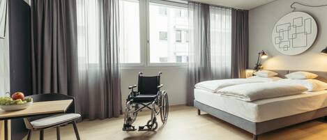 A modern hotel room with a wheelchair, a bed, a desk with a bowl of fruit, and a large window with curtains.