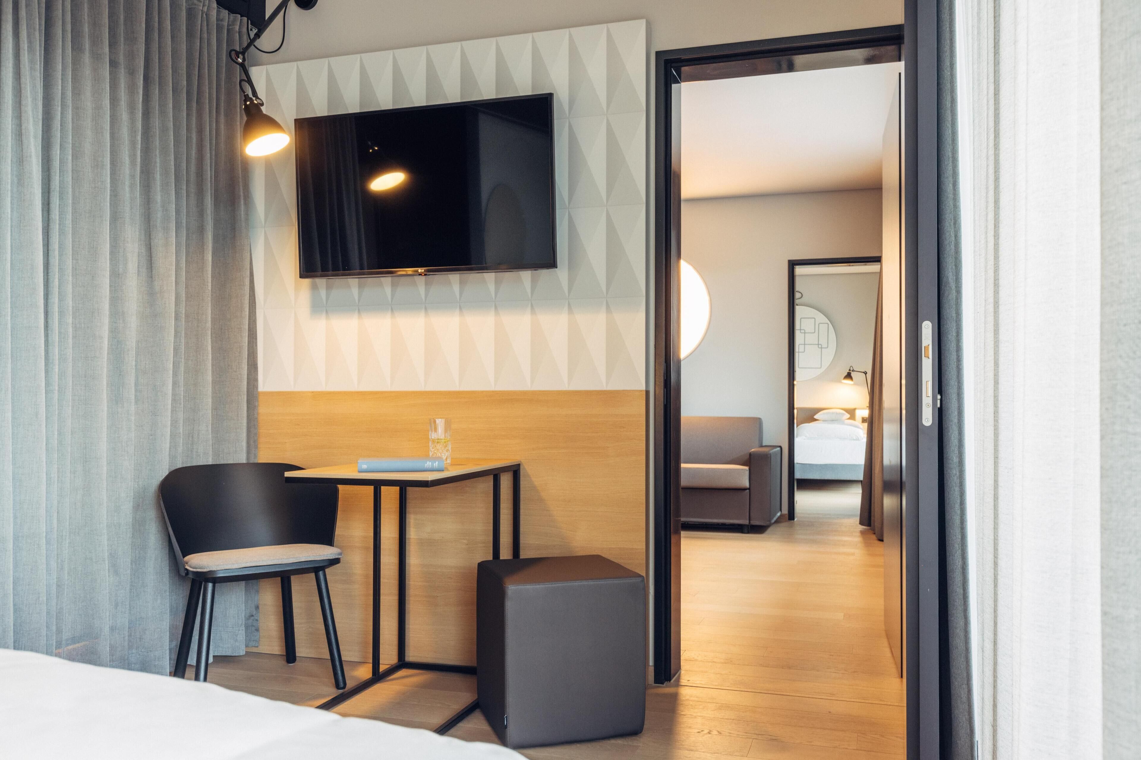 Habitación de hotel con cama, escritorio con silla, televisión montada en la pared y una pequeña mesa con un vaso.