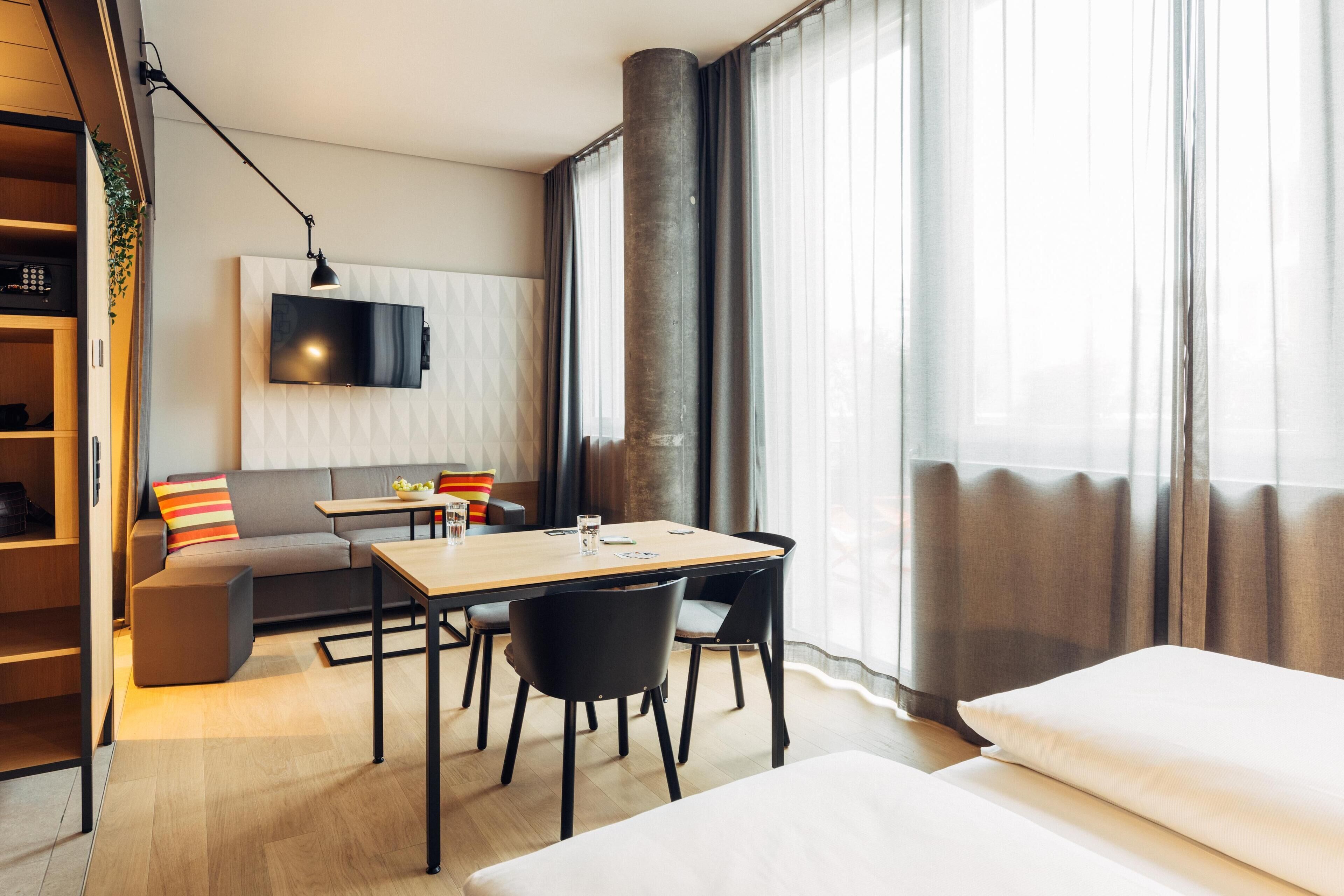 Et moderne hotelværelse med trægulv, sofa, spisebord med stole, fjernsyn ophængt på væggen og store vinduer med gardiner.
