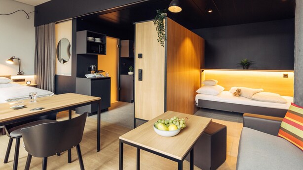 Una habitación de hotel moderna con una mesa de comedor de madera, un sofá, una pequeña cocina y una cama con mesita de noche.