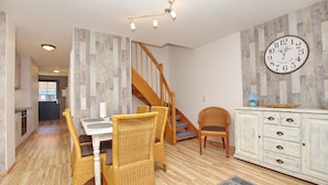 Un coin repas avec des chaises en osier, une table blanche, un escalier en bois et une horloge au mur.