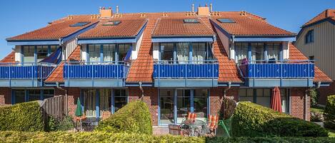 A two-story building with a red-tiled roof, balconies with blue railings, and a patio area with outdoor furniture.