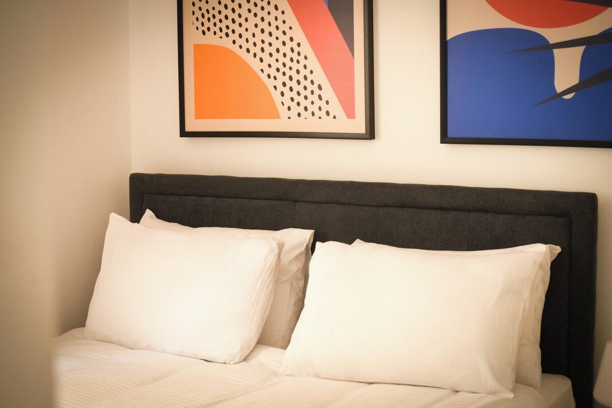 Una cama con dos almohadas y un cabecero frente a una pared con dos cuadros abstractos enmarcados.