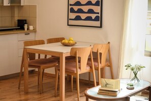 A dining area with a wooden table and chairs, a bowl of fruit, a framed artwork, and a small table with a book and flowers.