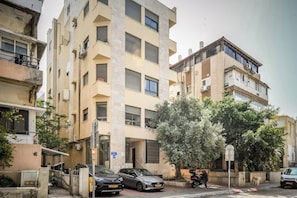 A multi-story building with balconies, parked cars, and a tree in front.