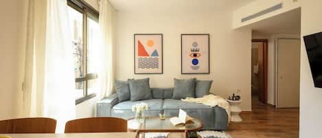 A living room with a sofa, a coffee table, and a dining table with a fruit bowl. There are framed pictures on the wall and a television mounted on the wall.